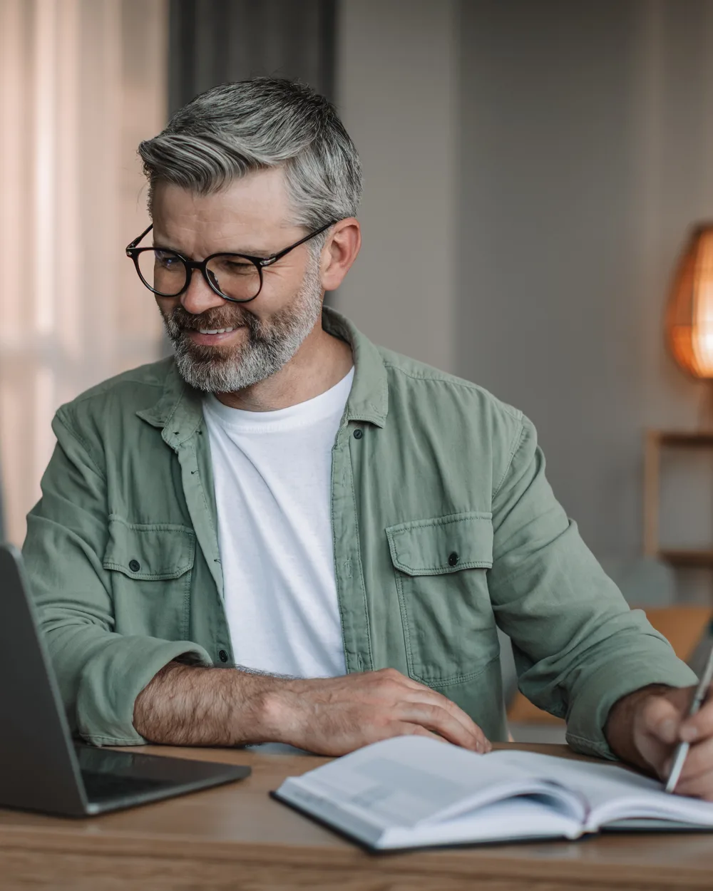 A mature professional smiles while taking notes during a live online coaching class at home.